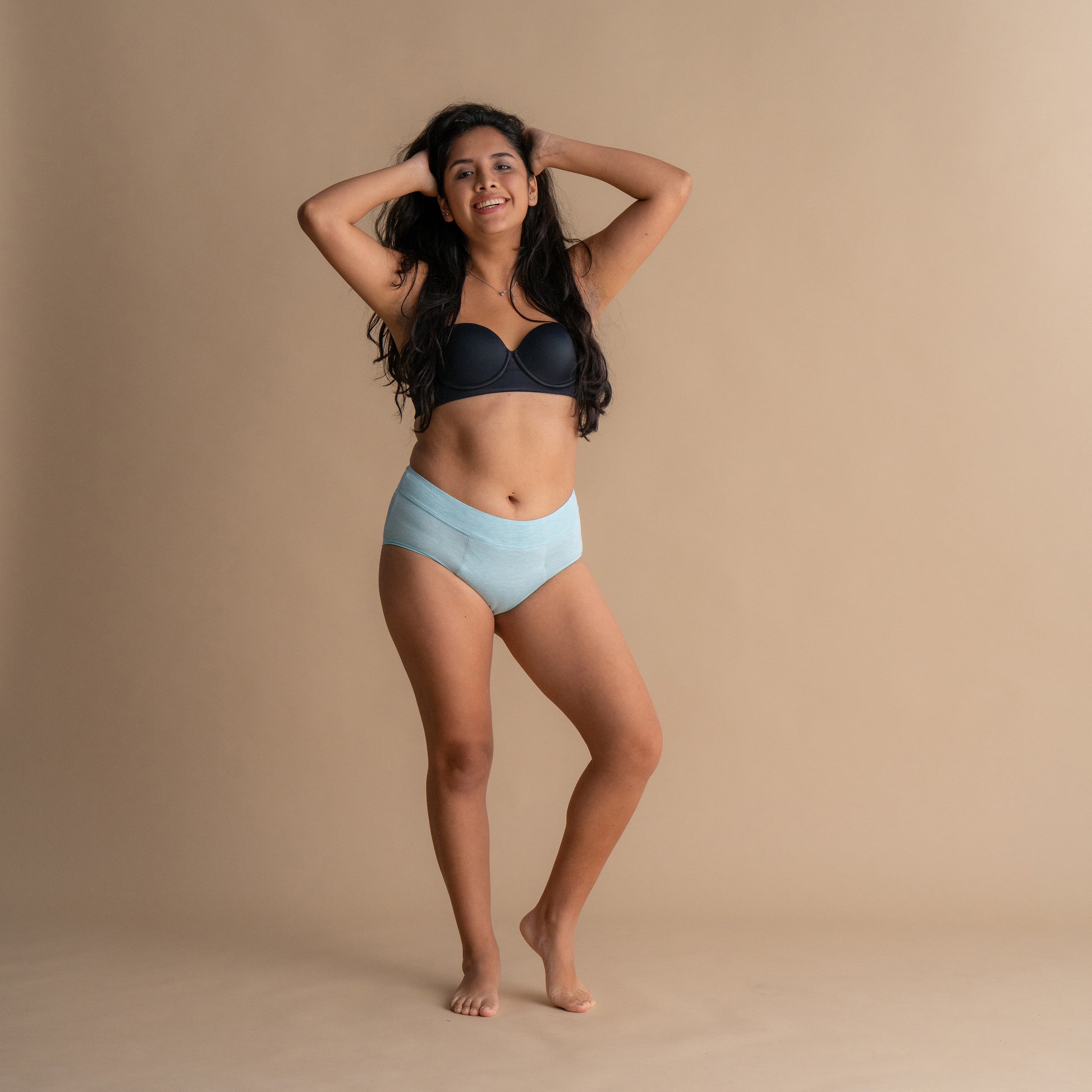 Nasmejan model v svetlo modrih Floena Leakproof Bamboo High Waist Brief, visoko pasne menstrualne hlačke, in črnem modrčku na bež ozadju