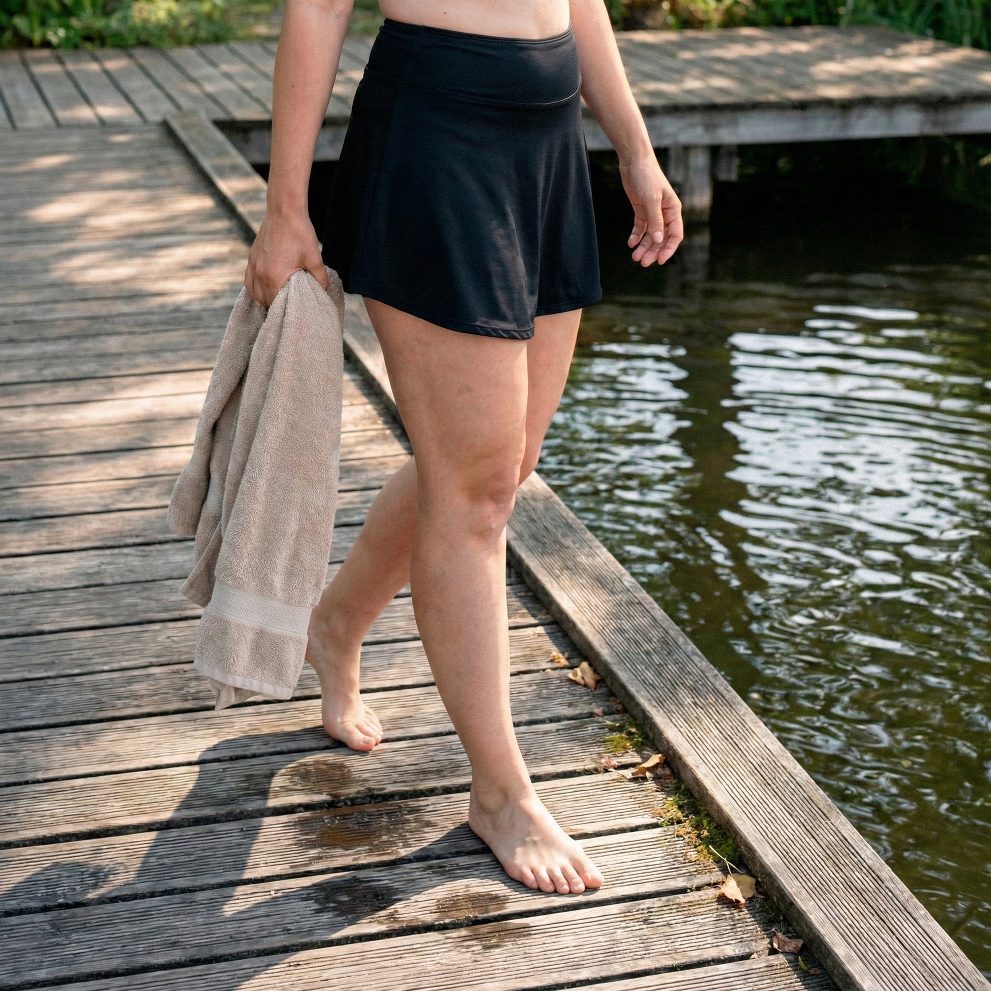 Model hodi bosa po lesenem pomolu ob vodi, drži brisačo in nosi črno Floena Leakproof Period Swim Skirt, menstrualno kopalno krilo