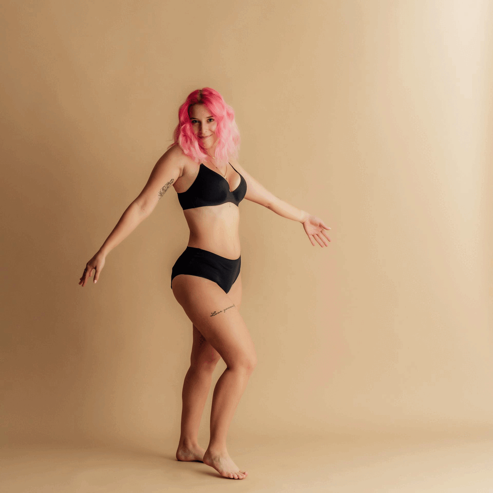 Ženska z roza lasmi, oblečena v črno Leakproof Period Underwear, se veselo pleše v studijskem okolju, izraža samozavest, udobje in telesno pozitivnost s sproščenim položajem.
