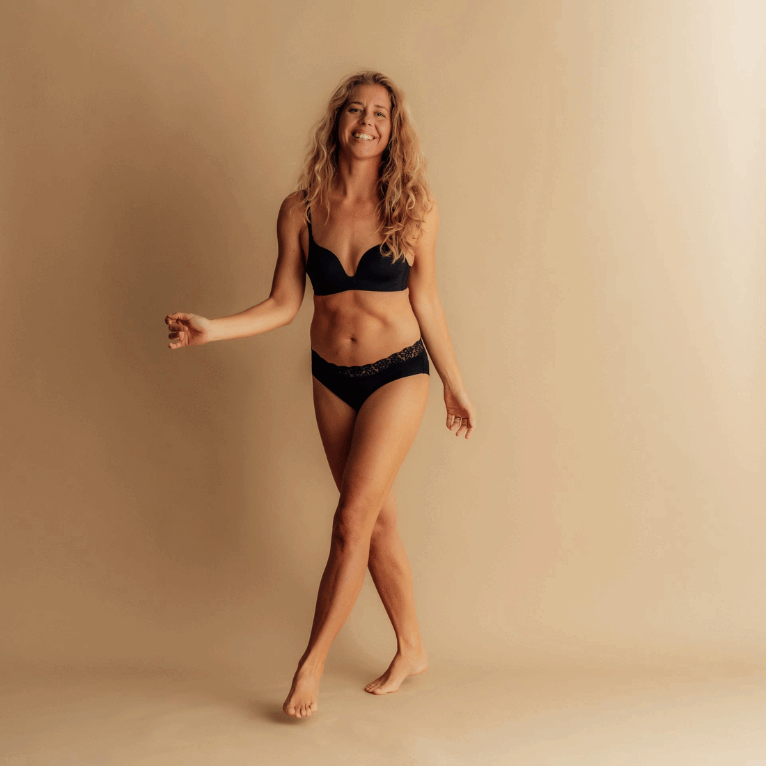 Ženska v črnih Leakproof Period Lace Brief, samozavestno razteza roke v sproščenem položaju pred nevtralno ozadje, ki predstavlja udobje in samozavest z zaščito proti puščanju.
